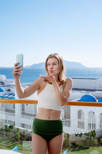 Portrait of young woman taking selfie against sea view from hotel balcony