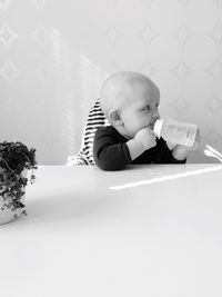 Baby boy drinking water while sitting at table against wall at home