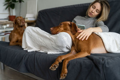 Full length of woman using mobile phone sitting with dogs sitting on sofa at home