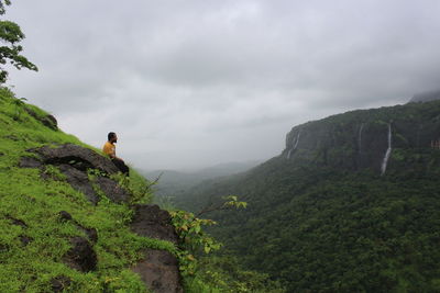 Mid adult man sitting on cliff against cloudy sky