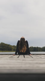 Rear view of woman sitting on pier over lake against sky