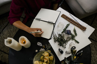 Midsection of woman writing in book by herbs and crystals on table