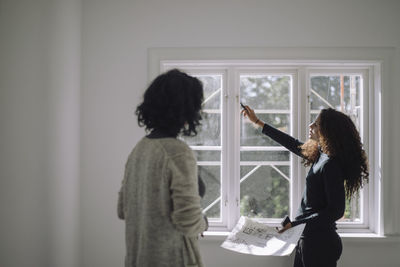 Female architect pointing at window while talking to client at under construction apartment