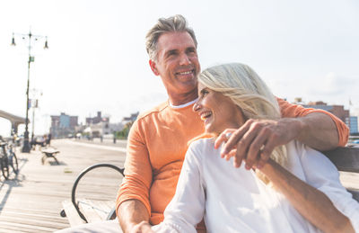 Happy couple sitting on bench against sky during sunny day