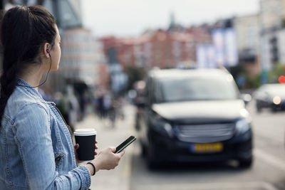 Side view of young woman holding mobile phone and coffee cup in city