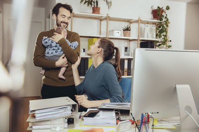Smiling mother at desk looking at father holding baby in home office