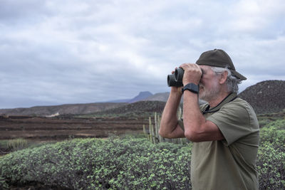 Side view of senior man looking through binocular on field against sky