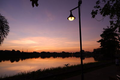 Silhouette street lights by lake against sky during sunset