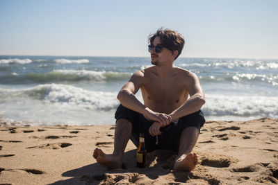 Shirtless young man sitting on beach against sky