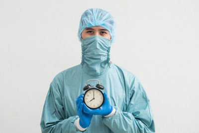 Portrait of surgeon holding alarm clock while standing against white background