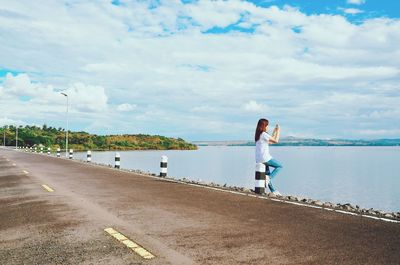 Woman sitting on bollard by road against sky