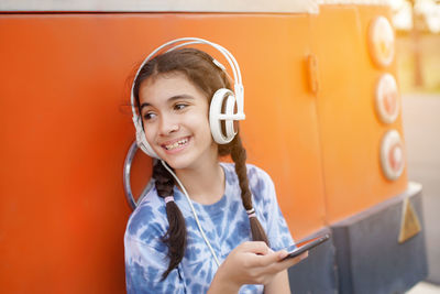 Smiling girl listening music while using smart phone against wall