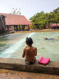 Rear view of shirtless boy sitting in swimming pool