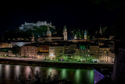 Illuminated buildings by river against sky at night