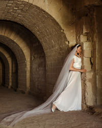 A sad elegant lady in white bride dress with a train languishes in the gloomy old castle