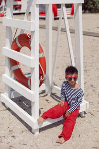 Child boy in striped clothes and red pants  standing on beach. white lifeguard tower, with a circle