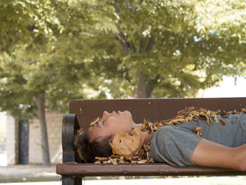Side view of teenager sleeping with fallen dry leaves on bench