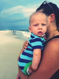 Side view of mother carrying son while standing at beach against cloudy sky