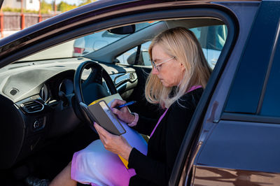 Middle aged business woman in glasses in car working with documents, mobile technology concept