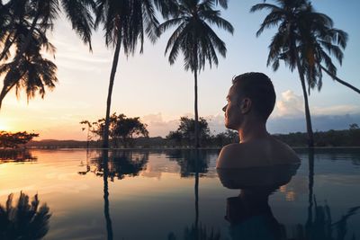 Young man watching sunset from swimming pool in the middle of coconut palm trees.