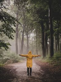 Rear view of woman with arms outstretched wearing raincoat while standing in forest