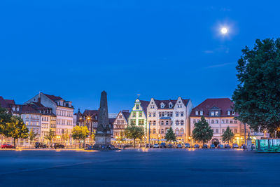 Illuminated buildings by street against blue sky at night