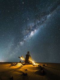 Rear view of man with illuminated string lights sitting on sand at desert against star field at night