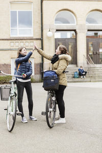 Happy female friends with bicycles high fiving outside school building