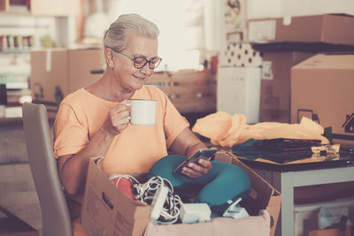 Smiling senior woman holding coffee cup using phone at home