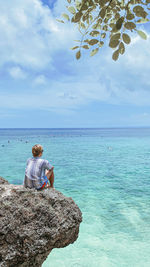 Rear view of woman sitting on rock by sea against sky