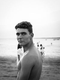 Portrait of shirtless young man standing at beach against sky