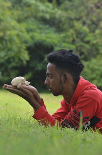 Side view of young man holding rock while lying on grassy land in forest