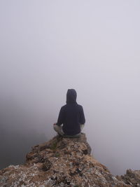 Rear view of person sitting at the edge of cliff during foggy weather