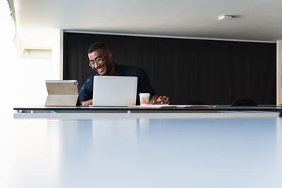 Positive african american adult male in formal clothes and eyeglasses sitting at table while having video call on tablet near netbook and documents with takeaway drink in bright office