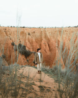 Young man in casual clothes with backpack and hat standing on path near arid canyon during trip in countryside