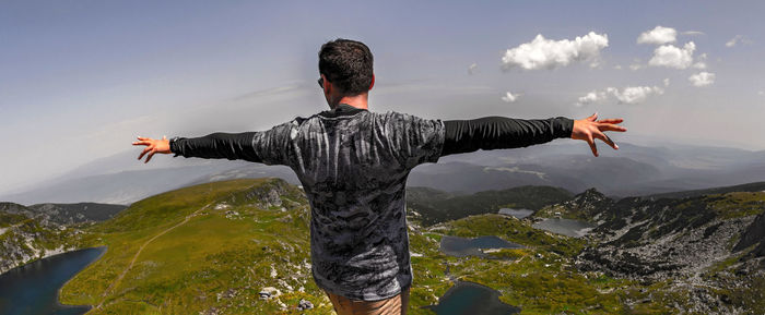Rear view of young man with arms outstretched standing on mountain against sky