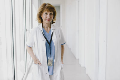 Portrait of senior female doctor standing with hands in pockets at hospital corridor