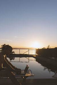 Side view of shirtless man using laptop while sitting by swimming pool against sky during sunset