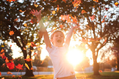 Portrait of happy boy with arms raised standing by falling leaves during autumn