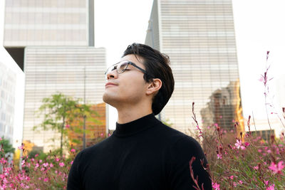 Handsome asian male in eyeglasses with eyes closed near bush with blossoming flowers on street on city
