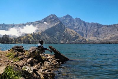 Side view of young woman crouching on rock at lakeshore against clear blue sky