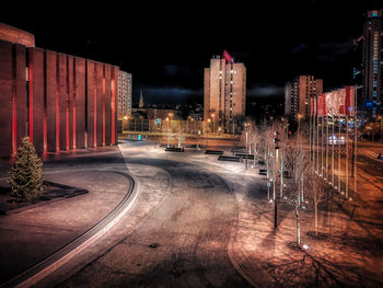 Illuminated road by buildings against sky at night