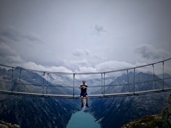 Man sitting on footbridge over mountains against sky