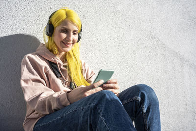 Portrait of a smiling young woman using mobile phone while sitting on wall