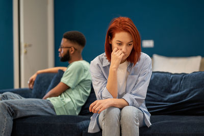 Upset diverse couple wife and husband sit separately on sofa at home not talking after quarrel