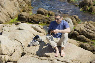 High angle view of mature man sitting on rock formation by stream during sunny day