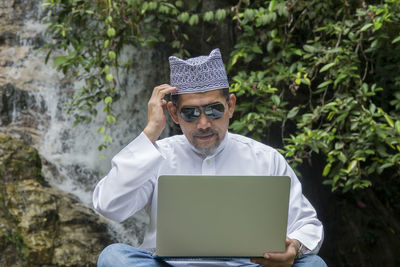 Mature man wearing sunglasses while using laptop against waterfall