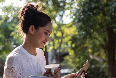 Smiling young woman holding cup using digital tablet while sitting in yard