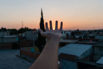 Cropped image of hand over city against sky during sunset