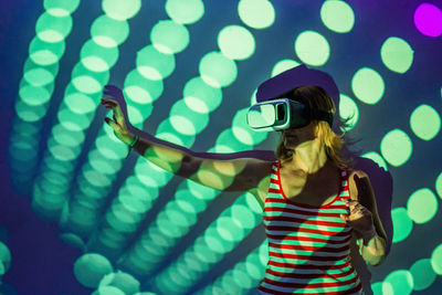 Unrecognizable young female in casual wear and vr headset getting new experience and touching virtual object in room with colorful projector illumination